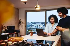 Womale and male standing next to the kitchen counter top, looking at a laptop. Smiling.
