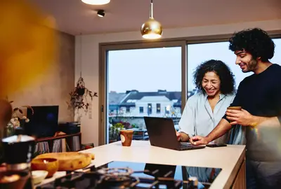 Womale and male standing next to the kitchen counter top, looking at a laptop. Smiling.
