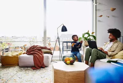 male sitting on couch with his laptop, smiling at a kid holding a notebook.
