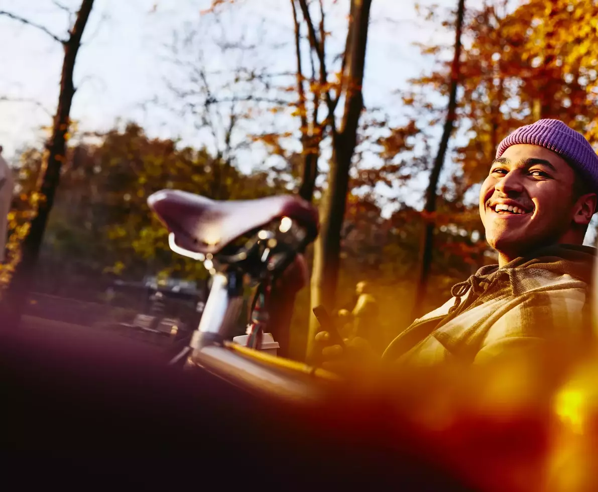 Smiling male sitting on a bench with his bike next to him, looking up while holding his phone.
