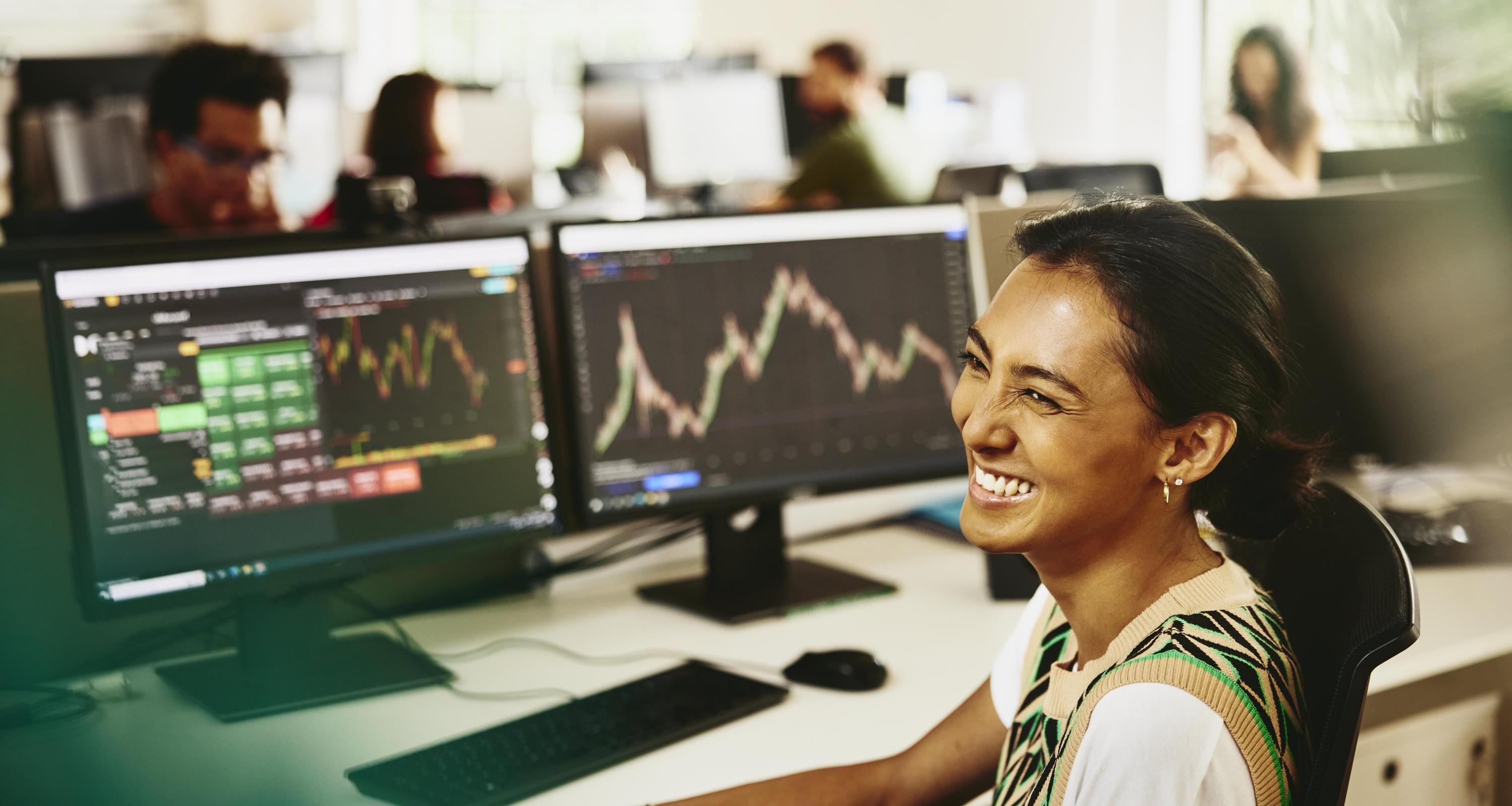 Woman smiling while sitting behind her desk, screens displaying financial information.
