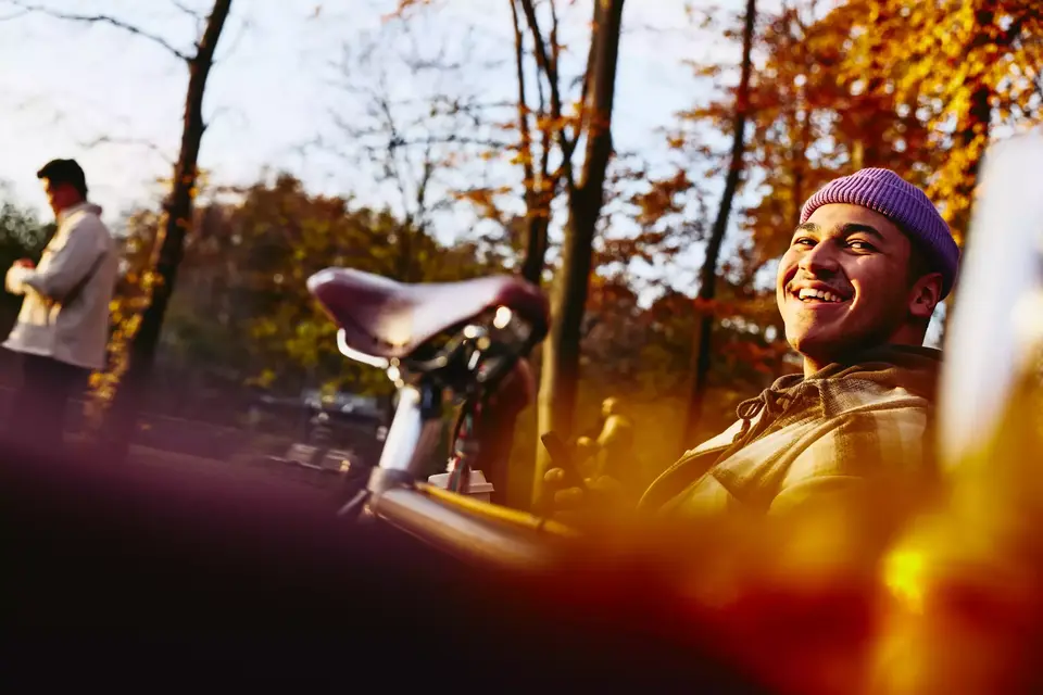 Smiling male sitting on a bench with his bike next to him, looking up while holding his phone.
