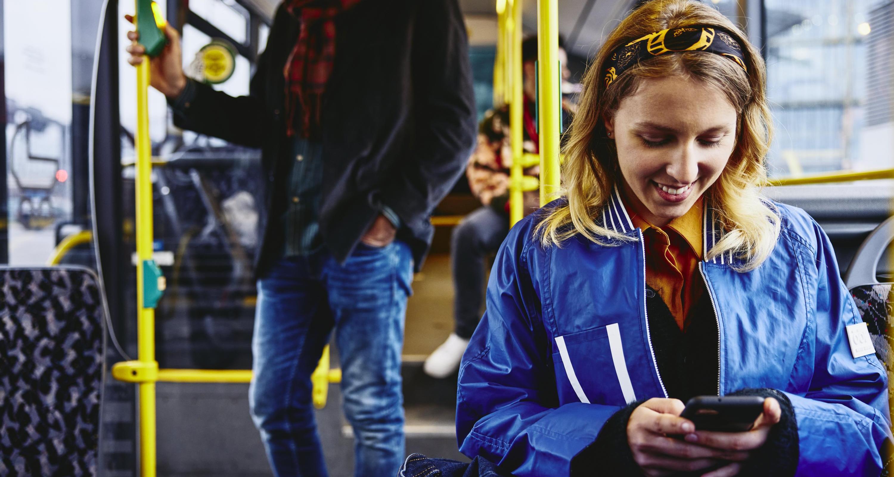 Smiling woman sitting in a bus, looking at her phone. Other people in the background.